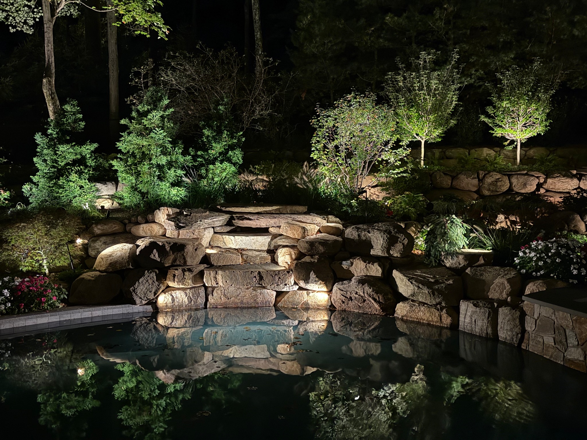 A stone waterfall and planting beds illuminated beside a pool